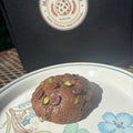 Chocolate pistachio cream cookie with chocolate chips and pistachio pieces on a floral plate in front of a Mannazello Treatz bakery box