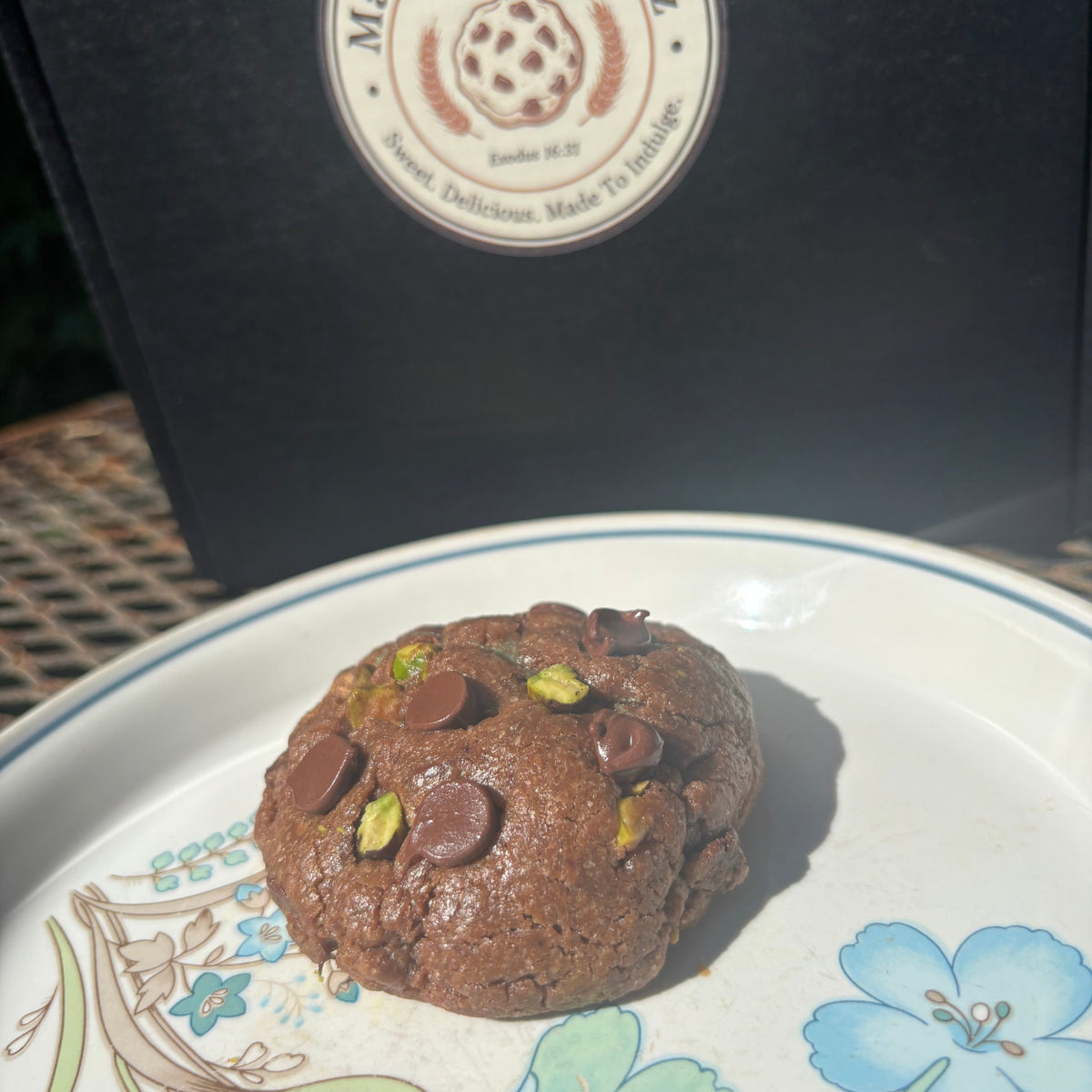 Chocolate pistachio cream cookie with chocolate chips and pistachio pieces on a floral plate in front of a Mannazello Treatz bakery box