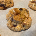 A close-up of a golden-brown cookie topped with cinnamon roll oreo pieces, cream cheese chips resting on parchment paper with other cookies blurred in the background.