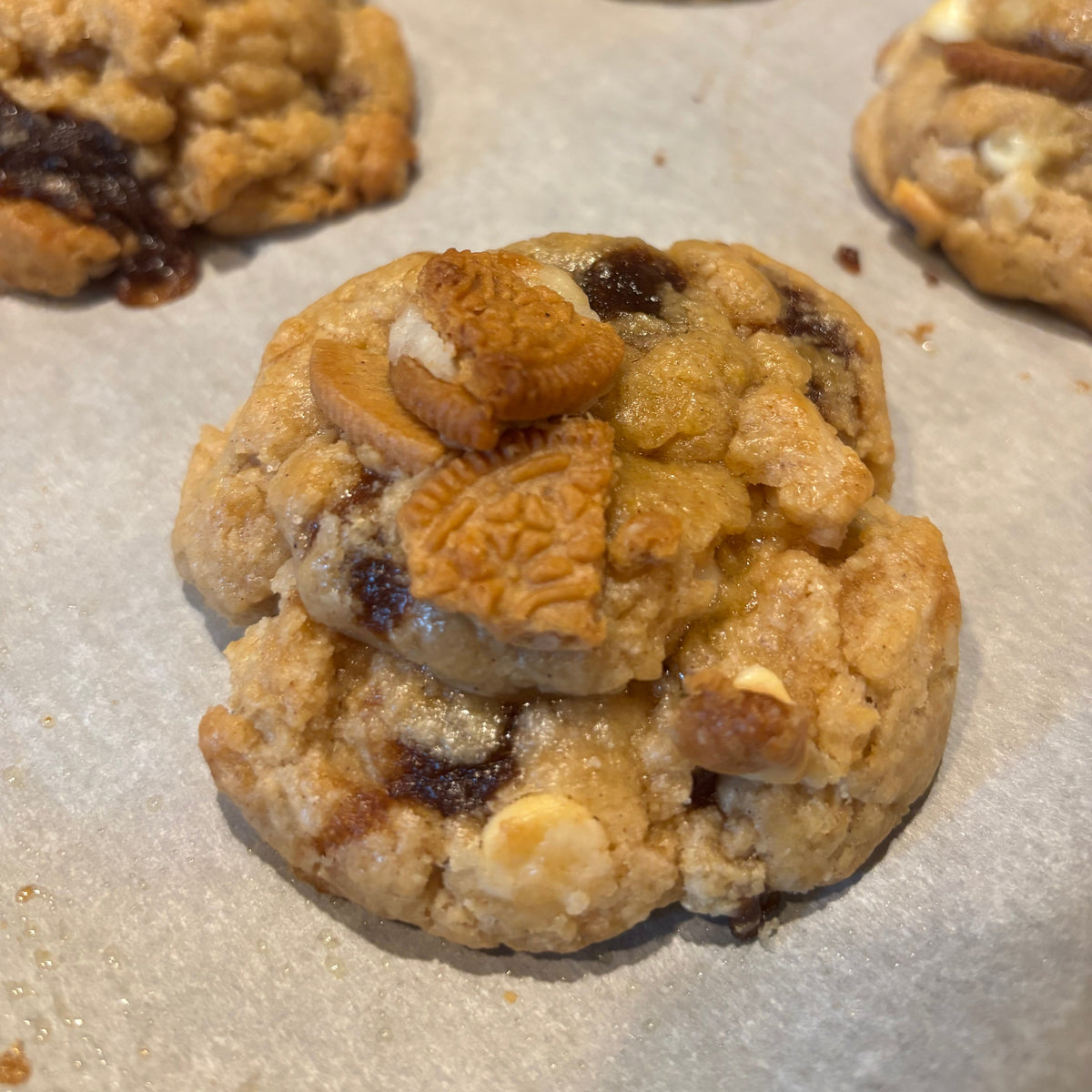 A close-up of a golden-brown cookie topped with cinnamon roll oreo pieces, cream cheese chips resting on parchment paper with other cookies blurred in the background.