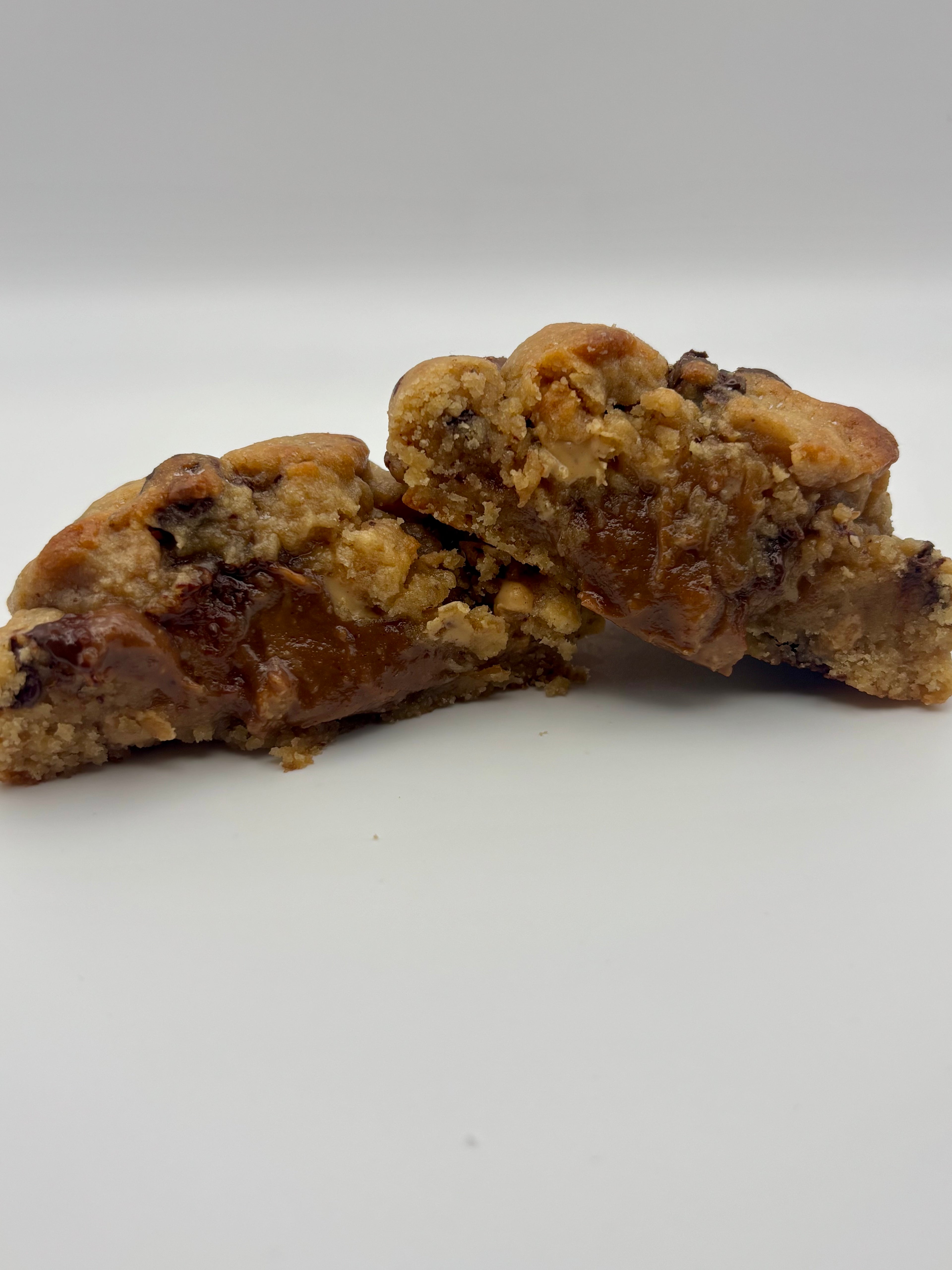 A close-up of a baked cookie broken in half to show the crumb texture, with peanut butter-chcolate filled center. Set against a plain light gray background.