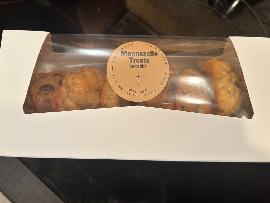 A white bakery box with a clear plastic window displaying golden-brown cookie treats inside. A round beige label in the center reads 'Mannazello Treatz Cookie Fight'.